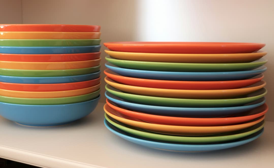 Dwa stosy kolorowych naczyń ceramicznych – z miskami po lewej stronie i ułożonymi warstwowo talerzami po prawej – tworzą pogodną i uporządkowaną kuchenną kompozycję.